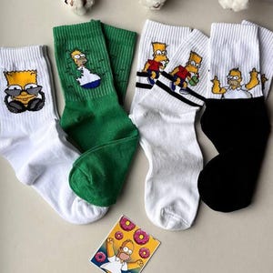 May include: Four pairs of socks featuring different Simpsons characters. The socks are white, green and black. One pair shows Homer Simpson with a donut.