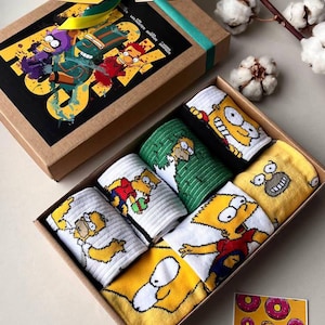 May include: A gift box filled with six pairs of colourful socks featuring cartoon characters from The Simpsons. The socks are in a brown box with a green ribbon and a tag that says "The Simpsons".