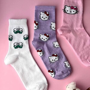May include: Three pairs of socks with different cartoon characters. One pair is white with green frogs, one pair is purple with pink Hello Kitty characters, and one pair is pink with a pink Hello Kitty character.