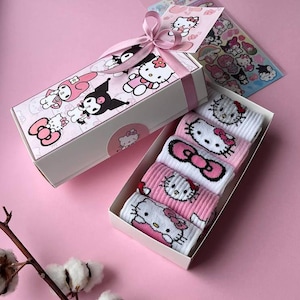 Puede incluir: Una caja de regalo rosa con una cinta blanca y personajes de Hello Kitty impresos en ella. La caja contiene un conjunto de cinco pares de calcetines de Hello Kitty en rosa y blanco.