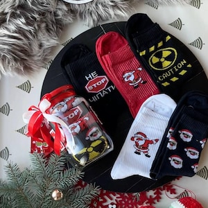 Könnte beinhalten: Eine Auswahl an Weihnachtssocken in Schwarz, Rot und Weiß. Einige Socken zeigen Santa Claus-Designs, während andere den Text "STOP" und ein Gefahrensymbol aufweisen. Die Socken werden mit einem dekorativen Glas und festlichen Akzenten präsentiert.