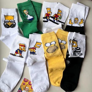 Könnte beinhalten: Eine Auswahl an Crew-Socken mit verschiedenen Designs von Charakteren aus der Zeichentrickserie Die Simpsons. Die Socken sind in Weiß, Grün, Gelb und Schwarz gehalten und zeigen Bart und Homer Simpson.