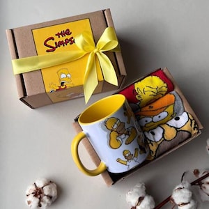 Könnte beinhalten: Ein Geschenkset mit einer weißen Tasse mit gelbem Griff und Homer-Simpson-Grafiken, Socken mit Zeichentrickfiguren und einer braunen Schachtel mit gelbem Band und "The Simpsons"-Logo.