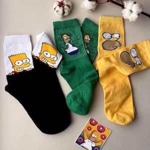 May include: Three pairs of socks with different coloured backgrounds and cartoon images of the Simpsons character Bart Simpson. The socks are green, yellow, and white with black accents. The socks are folded and arranged on a white surface.