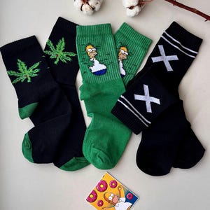 May include: A collection of colorful socks with various designs. Includes black socks with a green cannabis leaf, green socks with Homer Simpson, and black socks with white crosses. A small card with Homer Simpson and donuts is also present.