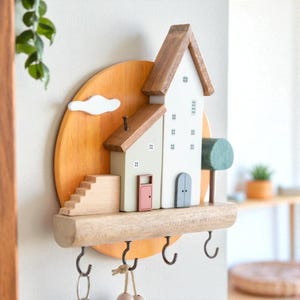 Puede incluir: Llavero de madera con forma de escena de pueblo en miniatura. Presenta una casa con tejado marrón, puerta rosa y puerta azul, sobre un fondo amarillo-naranja. Incluye una nube, escaleras, un árbol y tres ganchos para llaves.