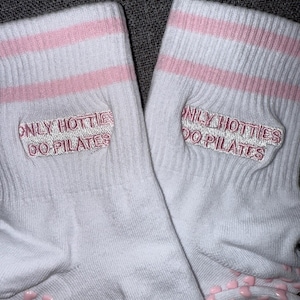 May include: A pair of white socks with pink stripes and pink heart-shaped grips on the bottom. The socks have the text "ONLY HOTTIES DO PILATES" embroidered on them in pink.