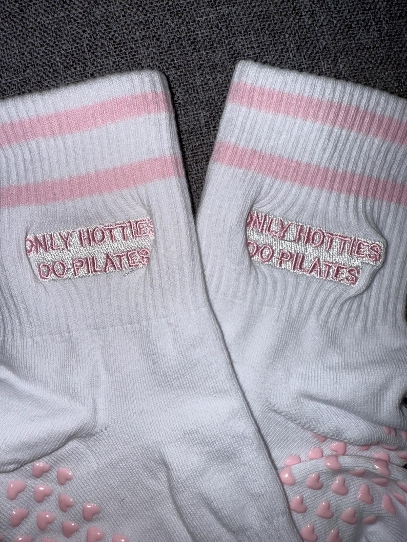 May include: A pair of white socks with pink stripes and pink heart-shaped grips on the bottom. The socks have the text "ONLY HOTTIES DO PILATES" embroidered on them in pink.