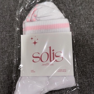 May include: White socks with pink stripes and pink dots on the bottom. The socks are packaged in a clear plastic bag with a white label that says "solis" in a stylized font. The label also says "Grip Socks" and "Shine with Every Move".