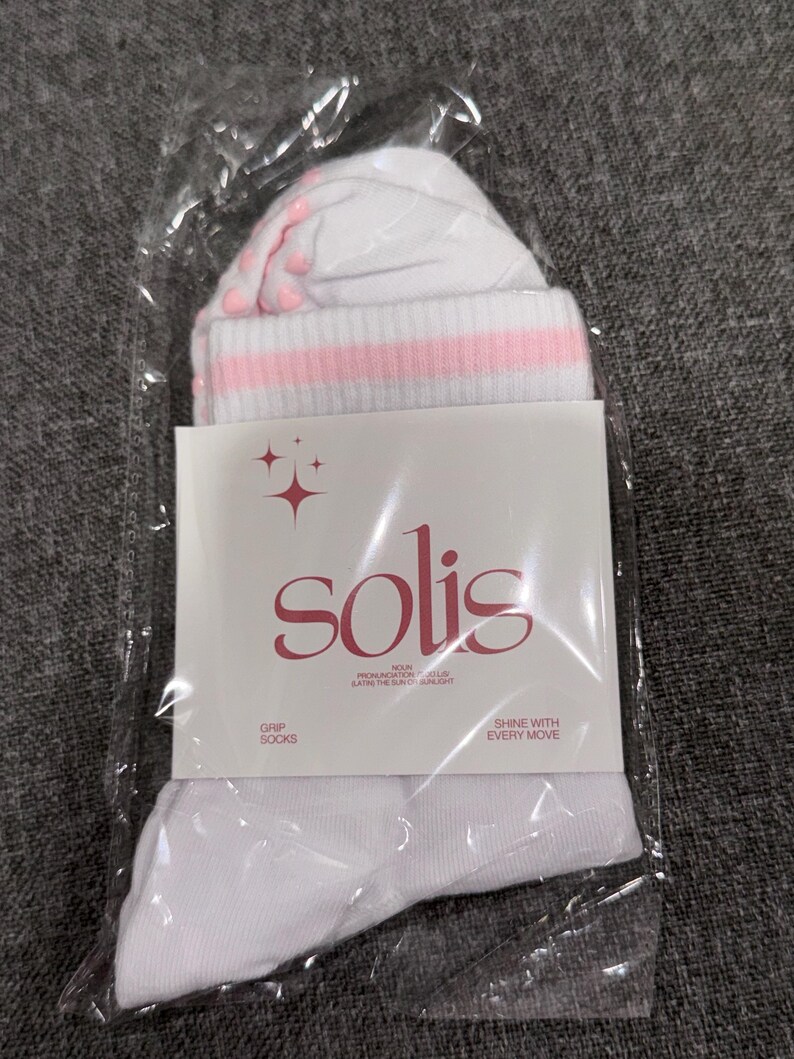 May include: White socks with pink stripes and pink dots on the bottom. The socks are packaged in a clear plastic bag with a white label that says "solis" in a stylized font. The label also says "Grip Socks" and "Shine with Every Move".