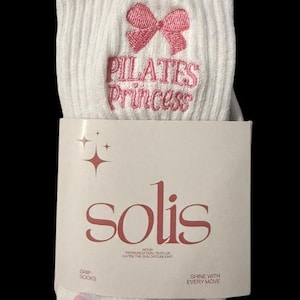 May include: White grip socks with pink non-slip dots on the bottom. The socks have a pink bow and the text "PILATES Princess" embroidered on the top. The socks are packaged with a white card that says "solis" in pink lettering.