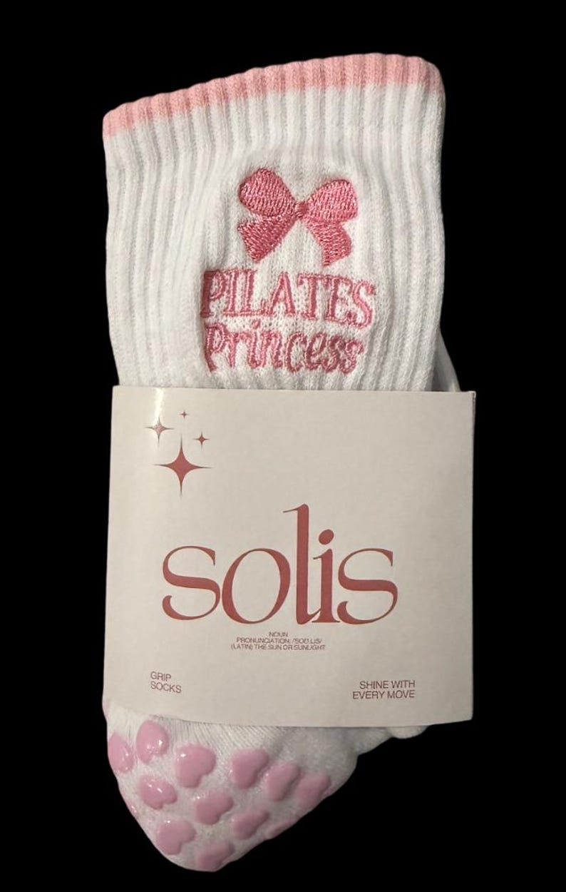 May include: White grip socks with pink non-slip dots on the bottom. The socks have a pink bow and the text "PILATES Princess" embroidered on the top. The socks are packaged with a white card that says "solis" in pink lettering.