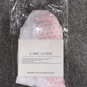May include: White socks with pink heart-shaped grips. The socks are packaged in a clear plastic bag with a white label that reads "CARE GUIDE" and includes instructions for washing and caring for the socks. The label also states "Material: 85% cotton, 10% polyester, 5% spandex".
