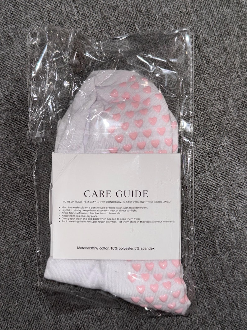 May include: White socks with pink heart-shaped grips. The socks are packaged in a clear plastic bag with a white label that reads "CARE GUIDE" and includes instructions for washing and caring for the socks. The label also states "Material: 85% cotton, 10% polyester, 5% spandex".