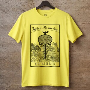 May include: A bright yellow short-sleeve t-shirt featuring a black graphic print. The design includes the text Björn Kornerup, Ars Longa Vita Brevis, and Ex Libris, surrounding an illustration of a signpost, a building, and trees.