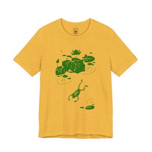 May include: Mustard yellow t-shirt featuring a green illustration of a frog leaping towards lily pads in a pond. The design includes lily pads, water ripples, and a water lily. The shirt has a crew neck and short sleeves.
