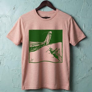 May include: A light pink t-shirt featuring a green graphic of two insects, possibly praying mantises or grasshoppers. The graphic is in a square frame and has a vintage, artistic style. The shirt is hanging on a wooden hanger.