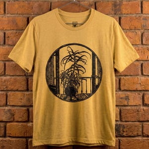 May include: Mustard yellow t-shirt featuring a black and white illustration of a potted plant inside a circular window. The design includes a balcony railing and a patterned rug. The shirt has short sleeves and a crew neck.