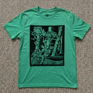 May include: A bright green short-sleeved t-shirt featuring a centered black graphic print of a stylized forest scene with a prominent tree in the foreground, tall trees in the background, and grassy ground. The initials E.H. appear in the corner.