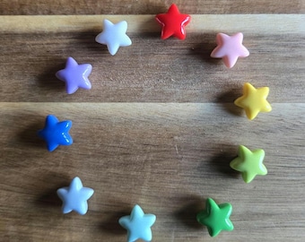 May include: A collection of nine colorful star-shaped beads arranged in a circle on a wooden surface. The beads are in various colors, including red, pink, yellow, green, blue, purple, and white. The beads are glossy and appear to be made of plastic.