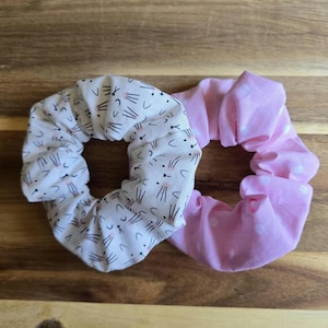May include: Two fabric scrunchies. One is white with a black cat face pattern. The other is pink with white polka dots. Both are circular and made of soft fabric. They are on a wooden surface.