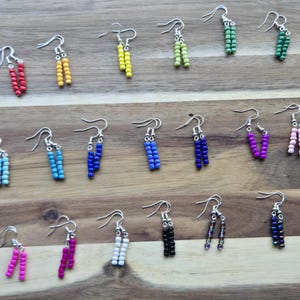 May include: A collection of beaded dangle earrings in various colors, including red, yellow, green, blue, purple, pink, white, and black. Each pair features a silver-tone hook and a vertical arrangement of small beads.
