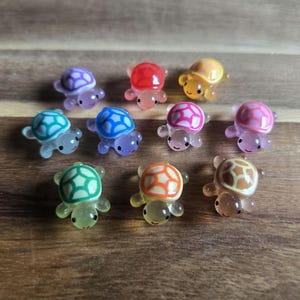 May include: A collection of ten miniature turtle figurines in various colors, including purple, red, yellow, blue, pink, green, and orange. Each turtle has a patterned shell and a smiling face, set against a wooden background.
