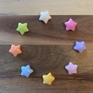 May include: A collection of eight star-shaped decorative objects in various pastel colors, including pink, blue, yellow, green, and orange. The stars are arranged in a circular pattern on a wooden surface.