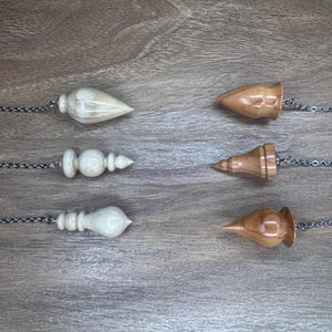 May include: Six wooden pendulums with silver chains are arranged on a wooden surface. Three are light-coloured, and three are a medium brown. Each pendulum has a unique shape, including teardrop, cone, and bulbous designs.
