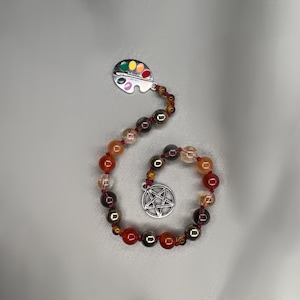 May include: A beaded necklace with a silver palette charm and a silver pentagram charm. The beads are in shades of red, orange, brown, and clear. The necklace is on a gray background.