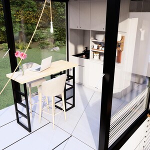 Op de afbeelding: Een modern, minimalistisch cabine-interieur met een kleine keuken en eethoek. De ruimte heeft een lichte houten tafel met twee witte stoelen, een laptop en een vaas met roze bloemen. Grote ramen bieden uitzicht op het groen.