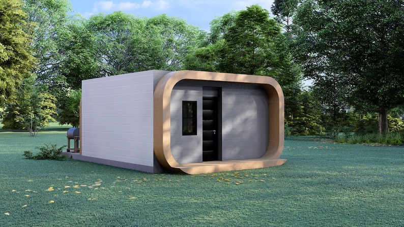 May include: A modern, minimalist cabin with a light gray exterior and a curved, wooden frame around the entrance. The cabin features a dark door and a small window, set against a backdrop of lush green trees and grass.