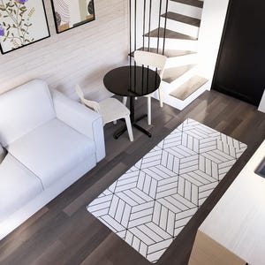 K&ouml;nnte beinhalten: Innenaufnahme eines modernen Wohnraums mit wei&szlig;em Sofa, gemustertem Teppich und kleinem schwarzen Tisch mit zwei St&uuml;hlen. Der K&uuml;chenbereich umfasst einen Herd und eine Sp&uuml;le. Kunstwerke h&auml;ngen an der Wand, und eine Treppe f&uuml;hrt nach oben.
