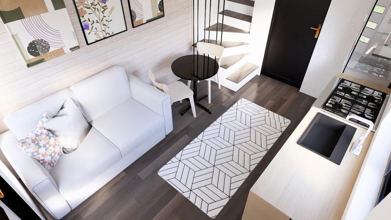 K&ouml;nnte beinhalten: Innenaufnahme eines modernen Wohnraums mit wei&szlig;em Sofa, gemustertem Teppich und kleinem schwarzen Tisch mit zwei St&uuml;hlen. Der K&uuml;chenbereich umfasst einen Herd und eine Sp&uuml;le. Kunstwerke h&auml;ngen an der Wand, und eine Treppe f&uuml;hrt nach oben.