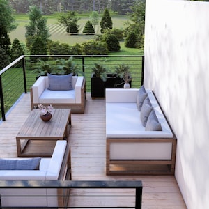 K&ouml;nnte beinhalten: Moderne Gartenm&ouml;bel mit Holzrahmen und wei&szlig;en Kissen. Das Set besteht aus zwei Sesseln, einem Sofa und einem Couchtisch. Die Terrasse hat ein Holzdeck und ein schwarzes Gel&auml;nder mit Blick auf einen gr&uuml;nen Rasen und B&auml;ume.