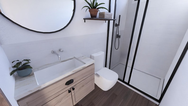 May include: A modern bathroom with a white toilet, a wooden vanity with a white sink, and a black-framed shower. A round black mirror hangs above the vanity. A small potted plant sits on the vanity and on a shelf above the toilet.