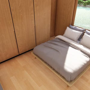 May include: A bedroom featuring light wood paneling and flooring. The bed has grey bedding and white pillows, positioned near a window. A small wooden bedside table with drawers and a plant is beside the bed. A large wooden wardrobe is on the left.