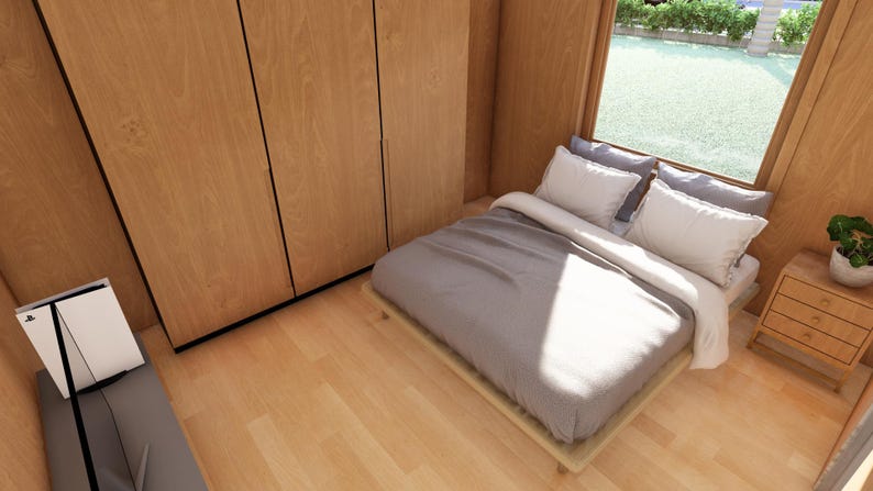 May include: A bedroom featuring light wood paneling and flooring. The bed has grey bedding and white pillows, positioned near a window. A small wooden bedside table with drawers and a plant is beside the bed. A large wooden wardrobe is on the left.