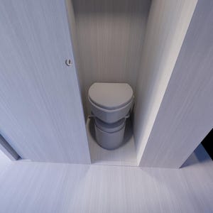 Op de afbeelding: Een lichtgrijze composttoilet staat in een kleine, wit ommuurde behuizing. Het toilet heeft een zitting en een lager gedeelte. De muren en vloer zijn lichtgrijs. Op de achtergrond is een bed met een blauw-wit gestreepte deken te zien.