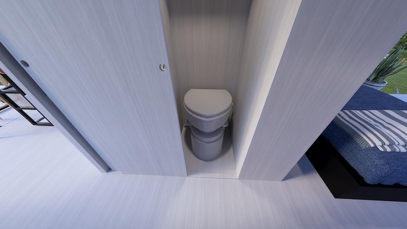 Op de afbeelding: Een lichtgrijze composttoilet staat in een kleine, wit ommuurde behuizing. Het toilet heeft een zitting en een lager gedeelte. De muren en vloer zijn lichtgrijs. Op de achtergrond is een bed met een blauw-wit gestreepte deken te zien.