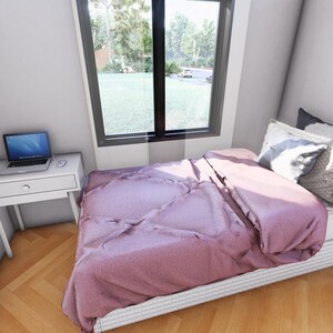 Puede incluir: Una escena de dormitorio con una cama cubierta con una colcha malva y almohadas. Tambi&eacute;n se ve un peque&ntilde;o escritorio blanco con una computadora port&aacute;til y una ventana con vistas a la vegetaci&oacute;n. La habitaci&oacute;n tiene piso de madera.