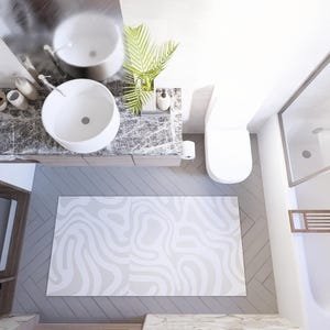 May include: An overhead view of a modern bathroom with a white sink, toilet, and shower. A gray and white patterned rug is on the floor. A washing machine is visible in a wooden cabinet. The bathroom has a minimalist design.
