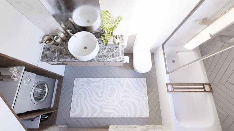 May include: An overhead view of a modern bathroom with a white sink, toilet, and shower. A gray and white patterned rug is on the floor. A washing machine is visible in a wooden cabinet. The bathroom has a minimalist design.