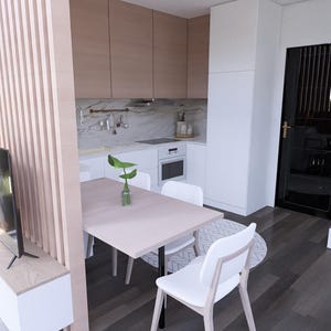May include: Interior view of a modern kitchen and dining space. It features light wood cabinets, a white countertop, and a dining table with white chairs. A large flat-screen TV is on a white console. The space is well-lit and contemporary.