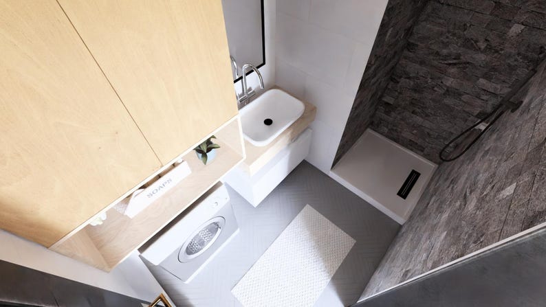 Op de afbeelding: Een moderne badkamer met een licht houten kast, een witte wastafel en een wasmachine. De doucheruimte heeft donkere, steenachtige tegels. Een wit tapijt ligt op de vloer.