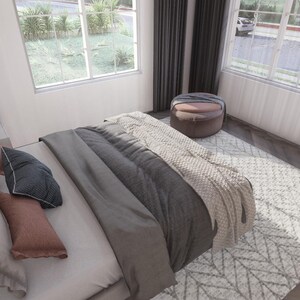 May include: A bedroom scene featuring a bed with gray and white bedding, including pillows and a throw blanket. A patterned rug sits on the wooden floor. A round ottoman and windows with curtains are also visible.