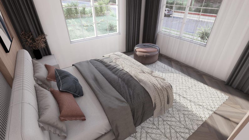 May include: A bedroom scene featuring a bed with gray and white bedding, including pillows and a throw blanket. A patterned rug sits on the wooden floor. A round ottoman and windows with curtains are also visible.
