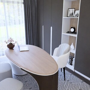 Op de afbeelding: Een moderne thuiskantoor met een ovale bureau van licht hout, een witte stoel en een ingebouwde boekenkast. De kamer heeft grijze gordijnen, een patroonkleed en een raam met uitzicht op groen. Een laptop en een vaas met bloemen staan op het bureau.