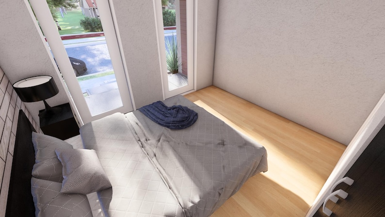May include: Bedroom interior with a bed, pillows, and a blanket. A black lamp sits on a black nightstand next to the bed. Large windows and a door provide natural light. The room has light-colored walls and hardwood floors.