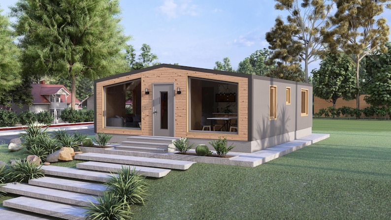 May include: A contemporary prefabricated house with a grey exterior and wooden features. Large windows reveal the interior, including a dining table. The house has a small front porch with steps and is surrounded by greenery.